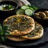 Freshly baked Manakish Za’atar flatbreads drizzled with olive oil, served with olives, cucumber slices, and mint on a dark slate platter