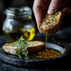 Hand dipping artisan bread into olive oil infused with Maison Herb & Spice No.3, with fresh herbs and oil jar in the background