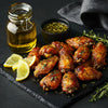 Golden roasted chicken wings garnished with herbs, served with lemon wedges, dipping sauce, and a bottle of olive oil on a black slate board