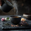 Hand pouring steaming herbal tea into a matte black teacup surrounded by dried flowers and loose tea leaves on a stone tray