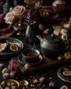 Elegant tea scene with steaming floral reserve tea in black ceramic teapot and cup, figs, roses, and dried botanicals on a dark luxury table setting