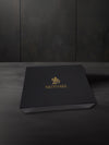 Angled shot of a closed Mediterra matte black gift box with gold logo, placed on a smooth dark surface against a plain dark gray wall background.