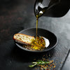 Elixir Gold No. 1 olive oil poured from t jar by Mediterra displayed on a luxury floor with Rosemary